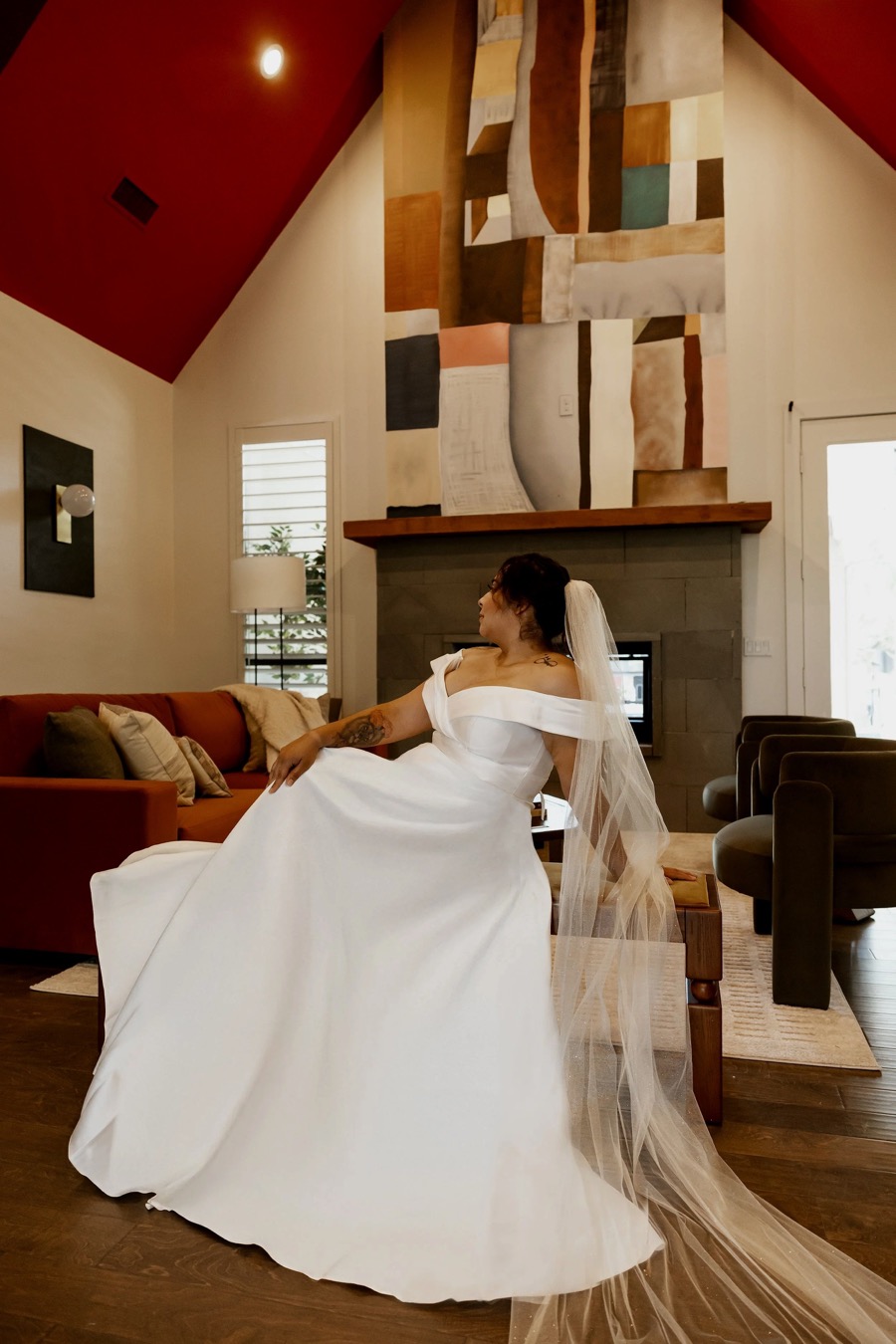 A woman dressed in a wedding gown and veil sitting in a living room, with a contemporary fireplace, abstract wall art, and modern furniture around her.