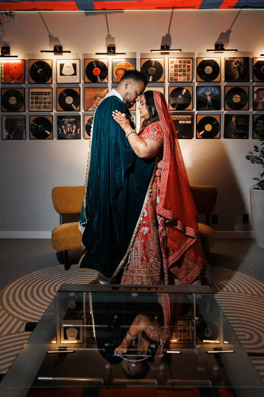 Indian bride in a red lehenga and groom in a teal velvet cape by the vinyl record wall in the Plus One Lounge at The Penrose House