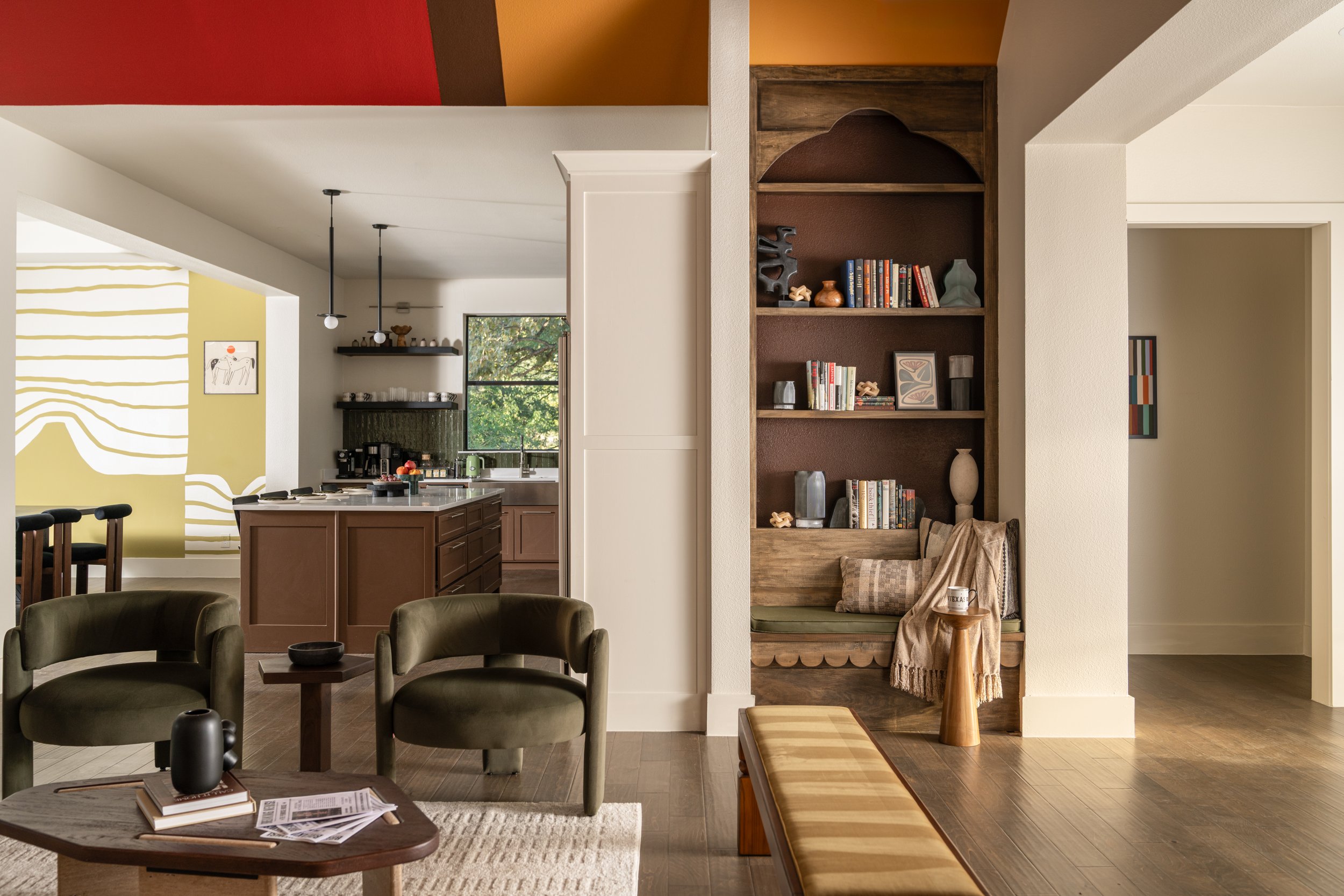 Living room open to kitchen with green armchairs, a wooden bench, and a built-in wooden bookshelf with decorative objects and books