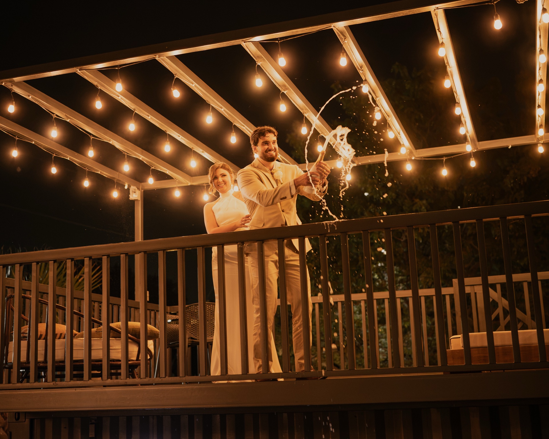 Bride and groom popping champagne on the upper bar deck beneath Edison bulb string lights at night