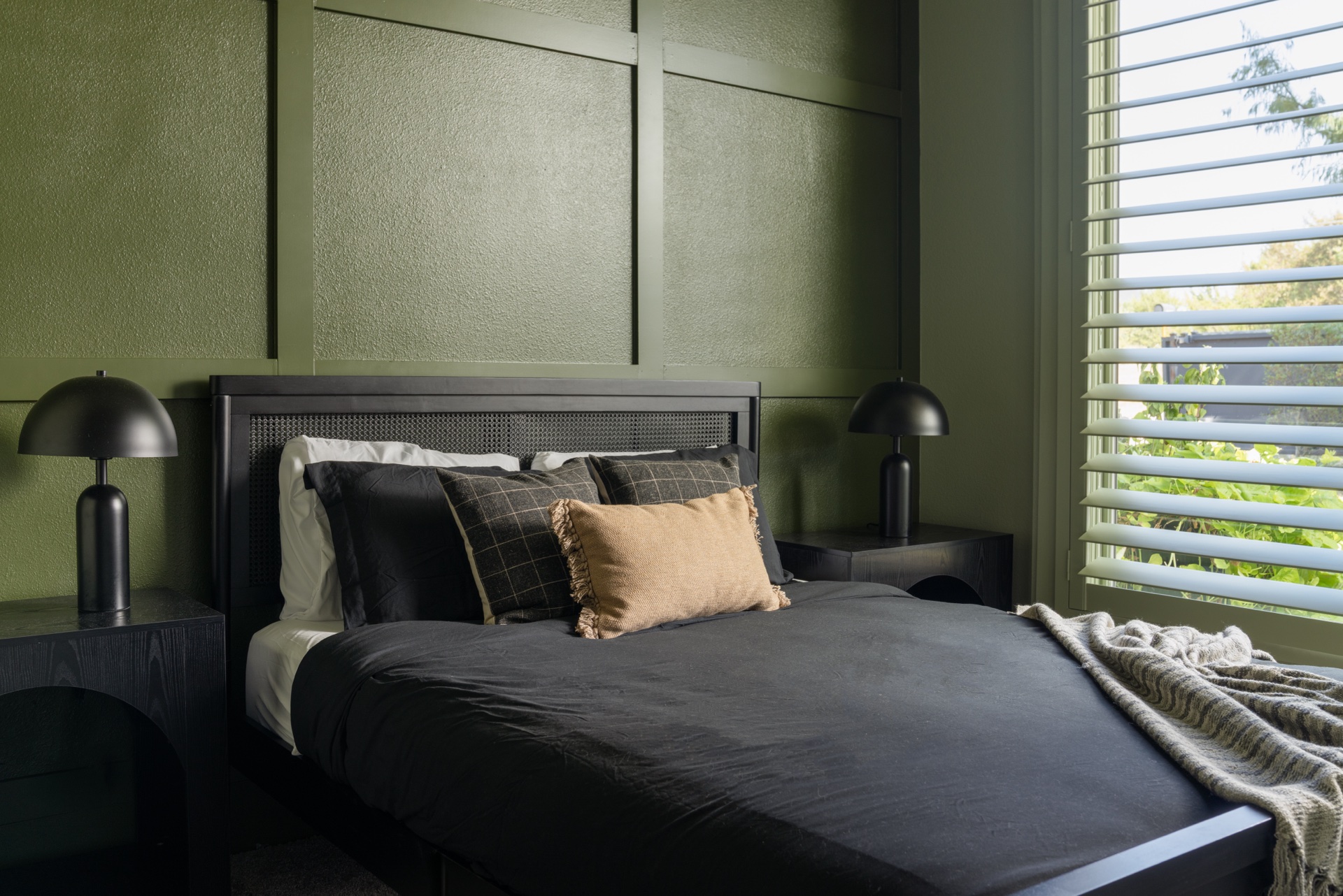 Bedroom with sage-green board-and-batten paneling, black bed with cane headboard, matching mushroom nightstand lamps and window with shutters at The Penrose House
