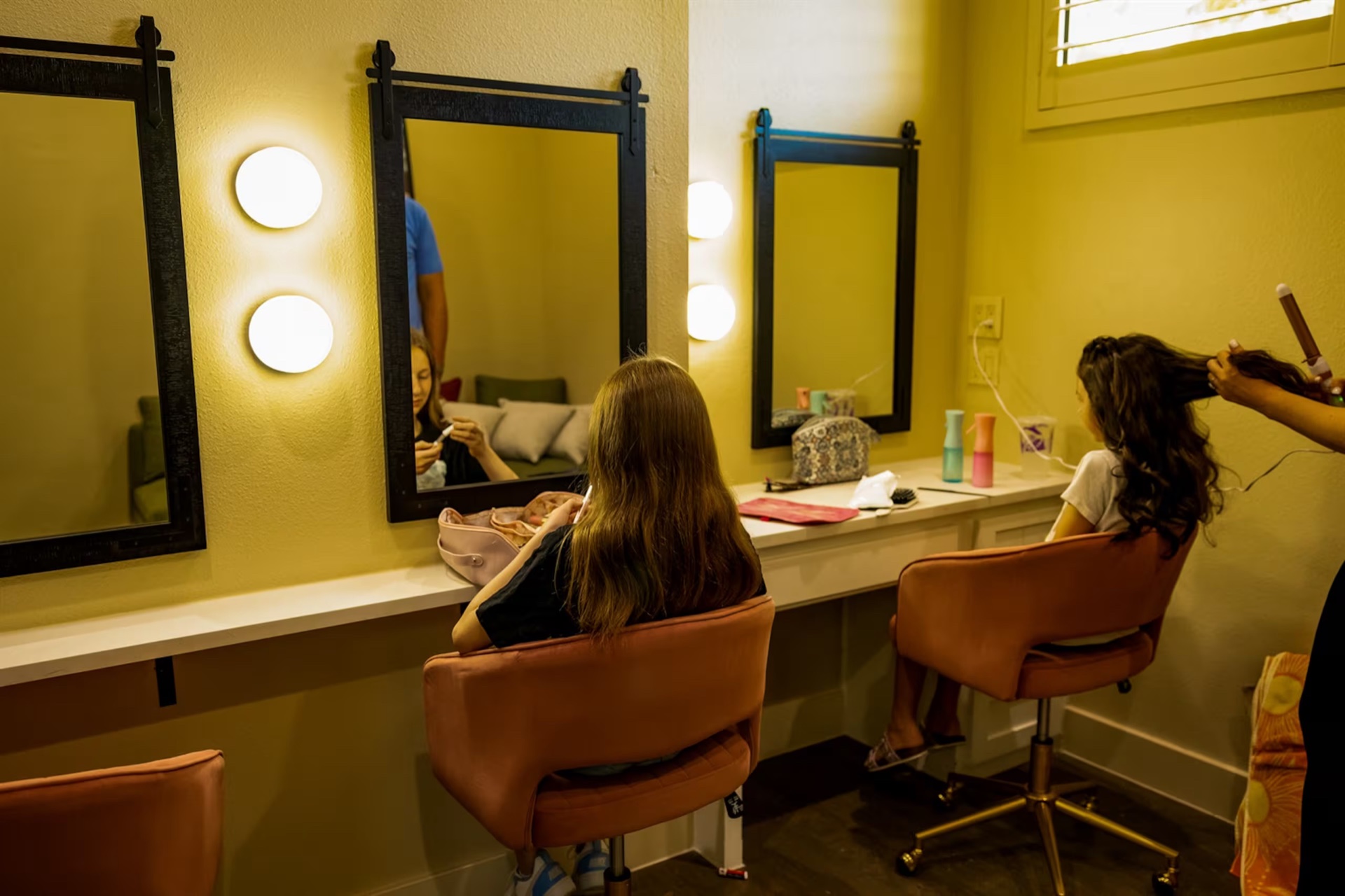 Two bridesmaids using the vanity stations in the Bridal Get Ready Room with illuminated mirrors, mustard walls and pink velvet chairs