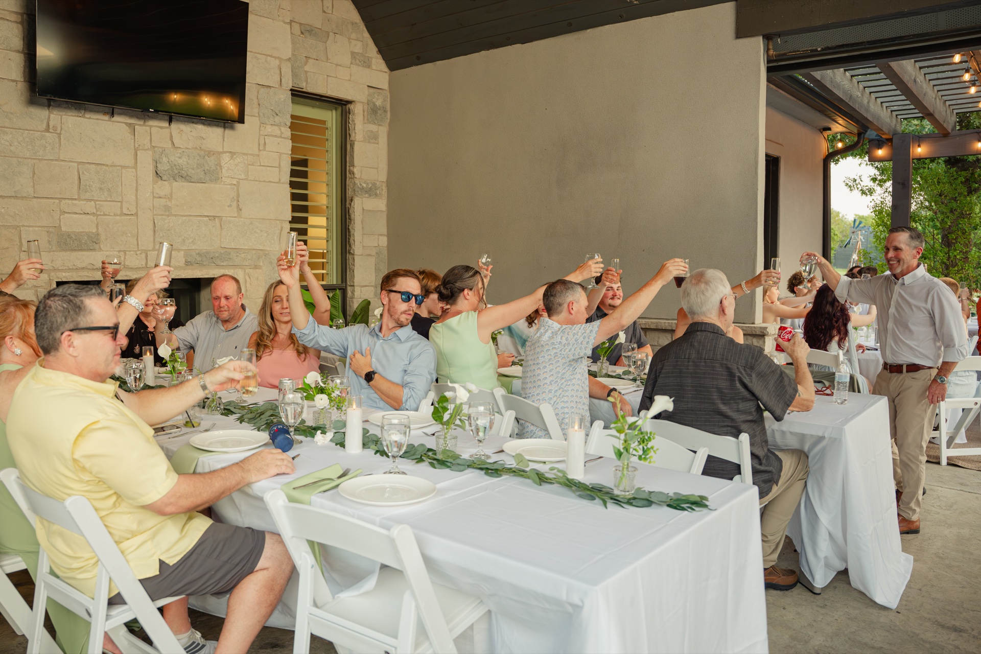 Wedding guests raising glasses for a toast at the long reception table on the covered dining patio with stone fireplace behind