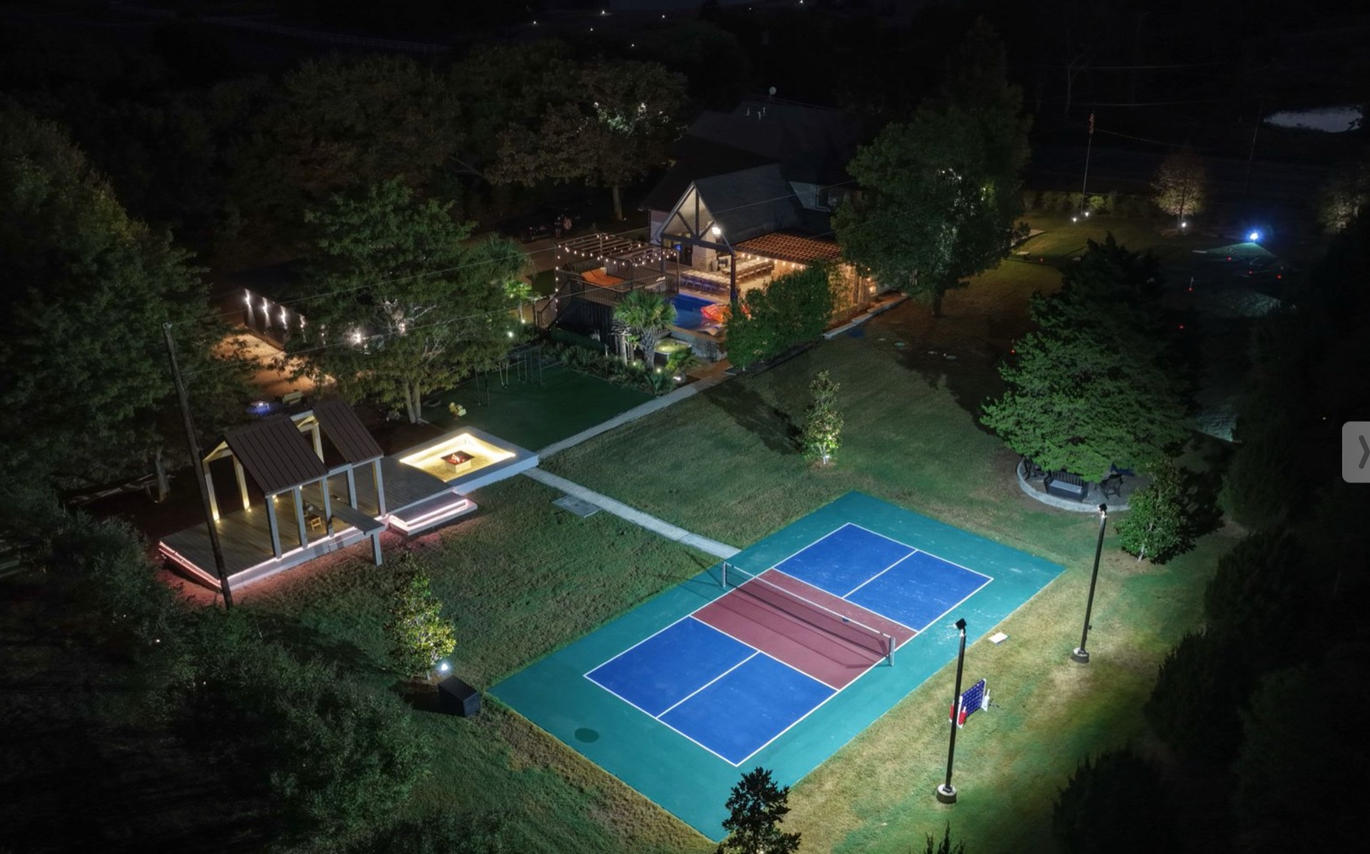 Aerial night view of the estate showing the lit pavilion, pickleball court, pool and string lights