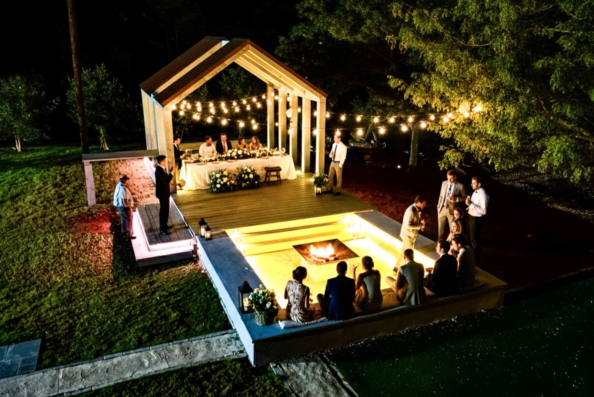 Aerial of the covered pavilion head table under string lights with firepit glowing and guests at dinner