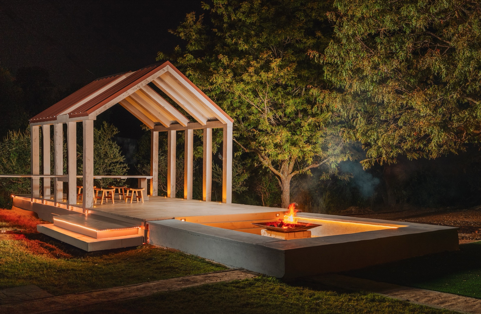 Covered wooden chapel pavilion with warm uplighting and open firepit at night