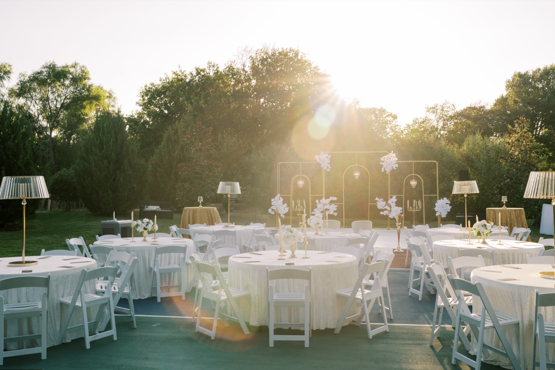 Round reception tables with white linens, gold candle lamps and floral arches with sunset backlight