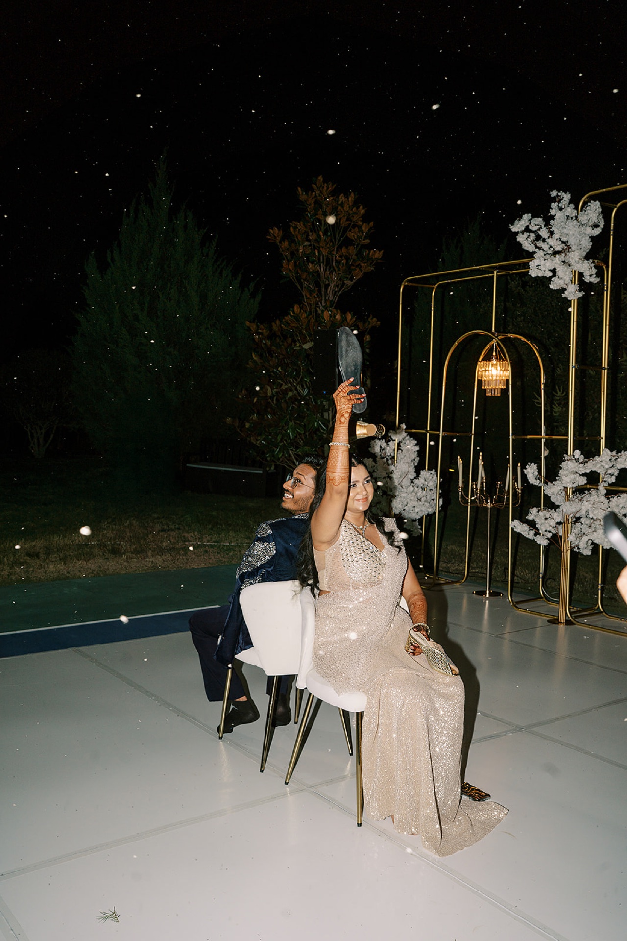 South Asian bride holding up a shoe during the shoe game as confetti falls, sparkle gown at night