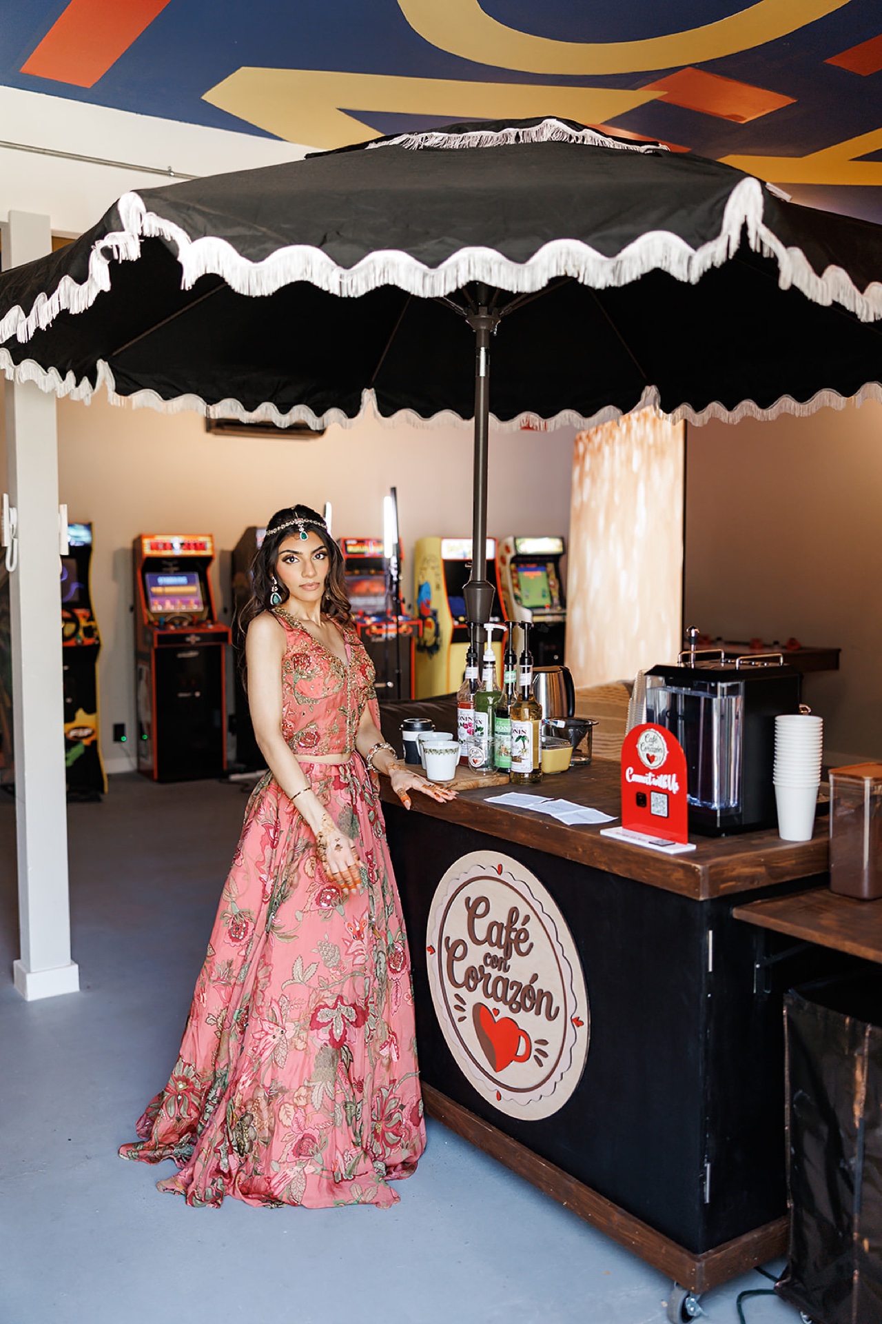 Guest in pink floral lehenga at the Café con Corazón coffee cart with arcade machines behind