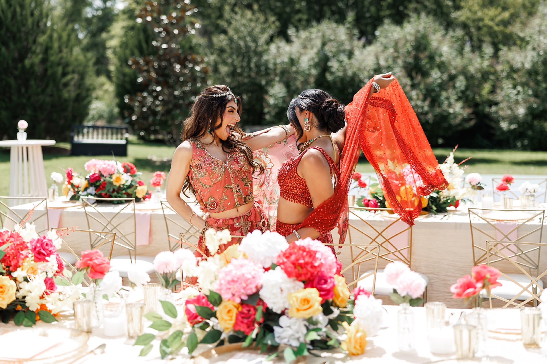 Two South Asian guests in orange and red lehengas laughing while draping a sequined dupatta at the reception