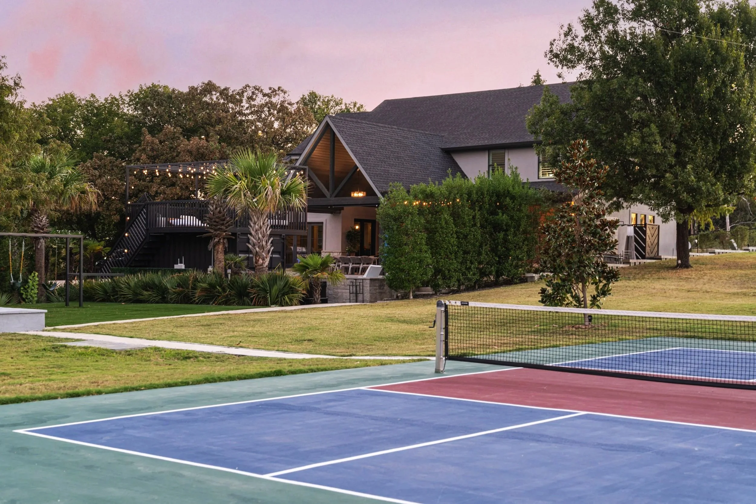 Pickleball court and estate at twilight with second-floor pergola lounge and string lights at The Penrose House