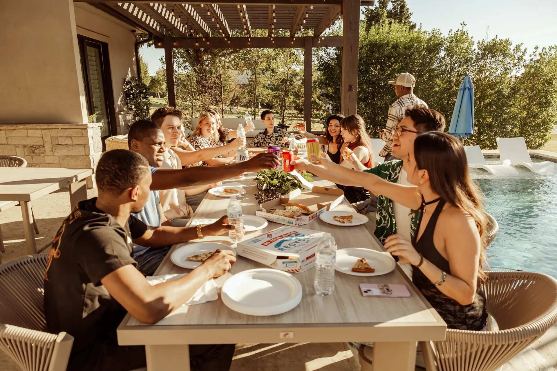 Group of diverse young friends celebrating with drinks and pizza at an outdoor poolside party.